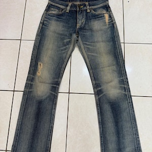 May include: Blue denim jeans with a faded wash and distressed details. The jeans have a classic five-pocket design, a button and zipper closure, and a straight leg. The denim has a worn appearance with light fading and some distressing on the front.