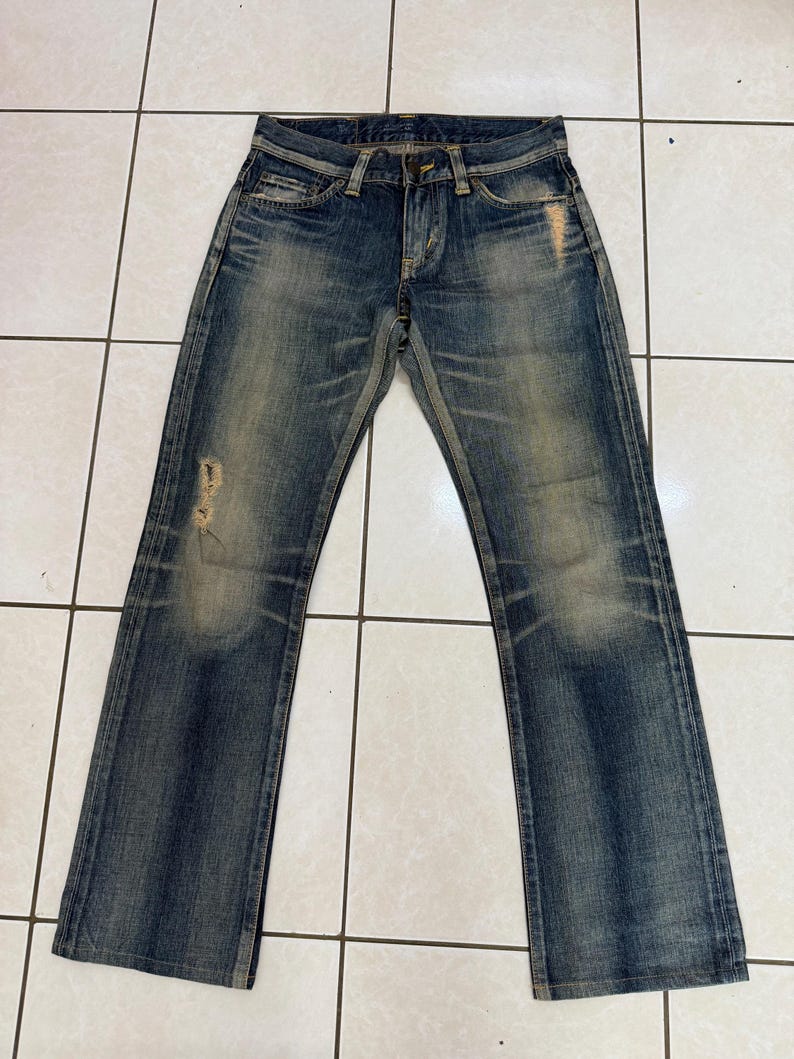 May include: Blue denim jeans with a faded wash and distressed details. The jeans have a classic five-pocket design, a button and zipper closure, and a straight leg. The denim has a worn appearance with light fading and some distressing on the front.