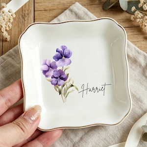 Könnte beinhalten: Eine weiße Keramik-Schmuckschale mit goldenem Rand und einem Blumendesign aus lila Stiefmütterchen. Der Name "Harriet" ist in schwarzer Schrift geschrieben. Die Schale ist quadratisch mit gewellten Kanten.