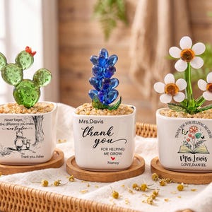 Op de afbeelding: Drie vierkante witte keramische plantenbakken op houten onderzetters, elk met een glazen plantensculptuur. Links een groene cactus, in het midden een blauwe bloem en rechts witte madeliefjes. Elke plantenbak is voorzien van aangepaste tekst en afbeeldingen.