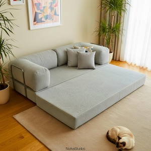 May include: A light gray modular sofa with a corduroy texture, featuring a chaise lounge extension. The sofa has several matching throw pillows in varying shades of gray. A beige rug and a sleeping cat are in the foreground.