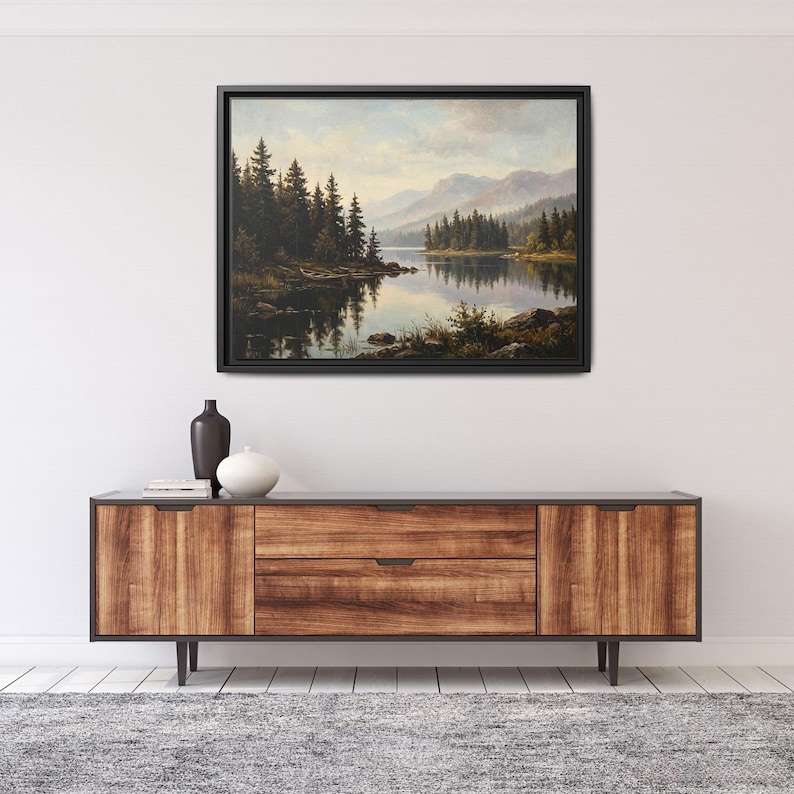 May include: A framed landscape painting depicting a calm lake surrounded by dense evergreen trees and distant mountains under a soft sky. The artwork is displayed in a thin black frame, mounted on a plain wall above a wooden sideboard.