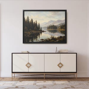 May include: A landscape painting in a black frame depicting a calm lake surrounded by dense pine forests and distant mountains under a soft sky. The artwork is mounted on a wall above a long, white four-door sideboard with gold hardware.