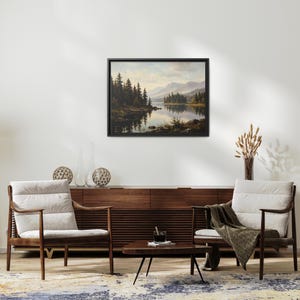May include: A framed landscape painting depicting a calm lake surrounded by dense evergreen trees and distant mountains under a soft sky. The artwork is displayed in a black frame on a plain wall above a wooden sideboard and two armchairs.