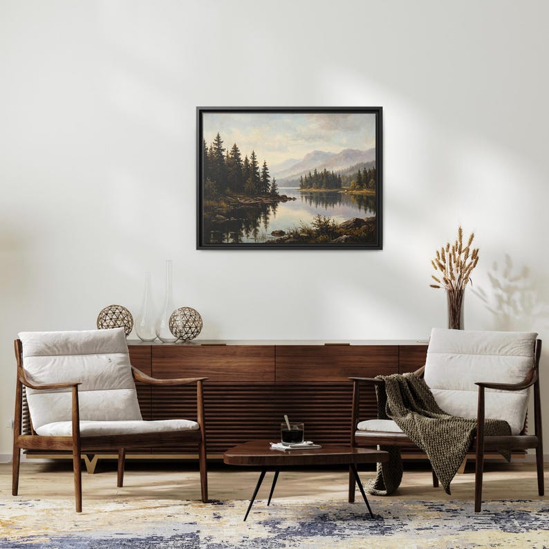 May include: A framed landscape painting depicting a calm lake surrounded by dense evergreen trees and distant mountains under a soft sky. The artwork is displayed in a black frame on a plain wall above a wooden sideboard and two armchairs.