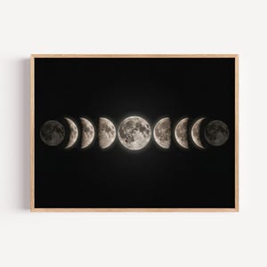 Puede incluir: Una impresión enmarcada que muestra las fases de la luna sobre un fondo negro. Las fases lunares se representan en una fila, pasando de la media luna a la luna llena y viceversa, con tonos grises y blancos. El marco es de color marrón claro.