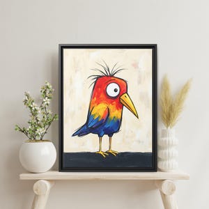 May include: A framed wall art print featuring a stylized illustration of a bird with a bright red, orange, and blue body, a yellow beak, and large white eyes with black pupils, set against a light beige background with a dark base.