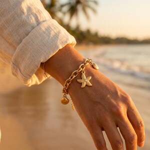 May include: Gold-coloured charm bracelet with a starfish, seashell, and chain link design. The bracelet is worn on a wrist, with a beach scene in the background. The bracelet is a nautical-themed accessory.