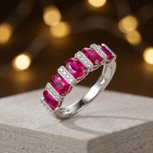 May include: A silver ring featuring oval-cut, vibrant red gemstones, alternating with sections of small, sparkling white stones. The ring is set on a neutral surface, with a blurred background of warm, golden lights.