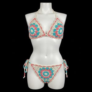 Peut inclure: Ensemble de bikini crocheté en crème, turquoise et rose, présenté sur un mannequin. Le haut de bikini a des bretelles de cou, et le bas a des côtés à nouer. Le haut et le bas présentent des motifs circulaires. Idéal pour la plage.