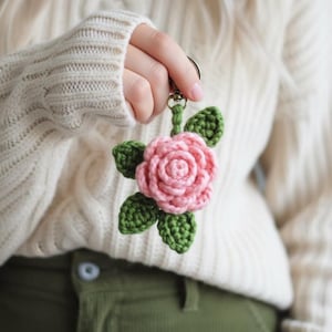 May include: A crochet keyring featuring a pink rose flower with multiple layered petals and four green leaves. The floral design is attached to a silver-toned metal split ring by a small green crocheted stem and a metal swivel clasp connector.