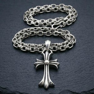 Puede incluir: Un collar plateado con un colgante de cruz. La cadena está hecha de eslabones entrelazados, y la cruz tiene un diseño detallado y ornamentado. El collar se muestra sobre una superficie oscura.