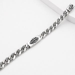 Op de afbeelding: Zilverkleurige gourmet armband met sierlijke kruis- en bloemmotieven. De armband heeft een rechthoekig middengedeelte met een gedetailleerd kruismotief en decoratieve schakels. Een stijlvol accessoire.