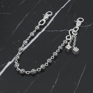 Può includere: Una catena per portafoglio color argento con una serie di maglie decorative a forma di sfera. La catena presenta due chiusure e due piccoli ciondoli a forma di dado appesi a una catena corta. I dadi hanno segni incisi.