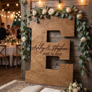 May include: A large wooden letter "E" with the names "Ashley & Stephan" and the date "June 18, 2027" engraved on it. The letter is decorated with white roses and greenery, with string lights above. A guest book and pen are in front.