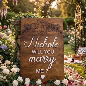 May include: A rectangular wooden sign with a dark brown finish featuring a black engraved floral design at the top. The sign displays white 3D lettering that reads Nichole WILL YOU marry ME ? with a small white heart icon at the bottom center.