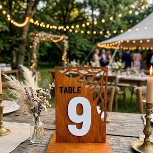 May include: A rectangular wooden table number sign featuring the text TABLE 9 in white and black lettering. The top portion of the sign has a laser-cut leaf pattern. The sign is mounted on a flat wooden base and stands upright on a rustic table.