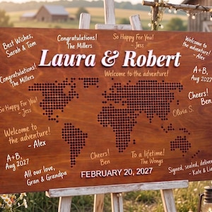 Könnte beinhalten: Ein hölzernes Hochzeitsschild auf einer Staffelei mit den Namen "Laura & Robert" in großen weißen Buchstaben. Das Schild zeigt ein Weltkarten-Design mit verschiedenen Glückwünschen und dem Datum 20. Februar 2027.