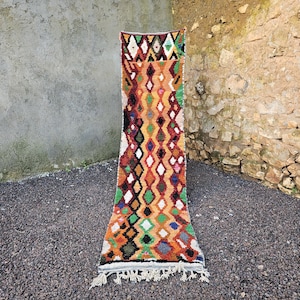 May include: A long, rectangular rug with a geometric pattern. The rug features a vibrant mix of colors, including orange, red, green, and white, arranged in a diamond pattern. The rug has a fringe at the bottom.