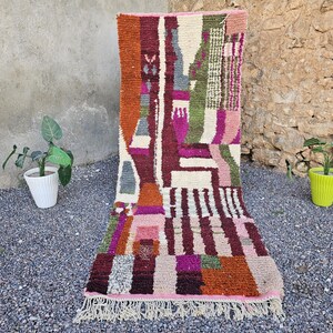 May include: A rectangular, colorful rug with an abstract design. The rug features a mix of geometric shapes and patterns in shades of orange, pink, green, and white. It has a fringed edge and is displayed outdoors.