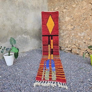Puede incluir: Una vibrante alfombra rectangular con fondo rojo y diseños geométricos. La alfombra presenta una forma de diamante amarillo, líneas negras y detalles azules y verdes. La alfombra tiene un borde con flecos y se exhibe al aire libre.