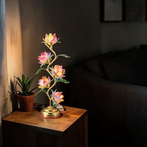 May include: A decorative table lamp with a floral design. The lamp has a gold-coloured base and stem, with four colourful lotus flower shades. The flowers are pink, yellow, and orange. The lamp is on a wooden table.