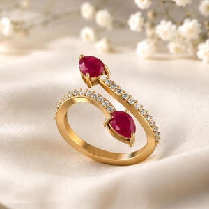 May include: A gold bypass ring featuring two pear-shaped red rubies at the open ends. The band is accented with a row of small, clear, round-cut white stones along the top and bottom edges, creating a sparkling, open-ended design.
