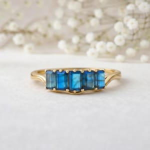 Puede incluir: Un anillo de oro con cinco piedras preciosas rectangulares de color azul vibrante. El anillo tiene un diseño delicado y curvado con una banda sutil y texturizada. Las piedras están dispuestas en una fila, creando una joya llamativa y elegante.