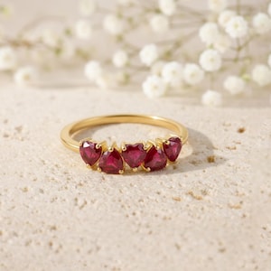 Può includere: Un anello d'oro con cinque pietre preziose rosse a forma di cuore. L'anello è posto su una superficie chiara e strutturata, con fiori bianchi sullo sfondo. Le pietre sono distanziate uniformemente lungo la fascia.