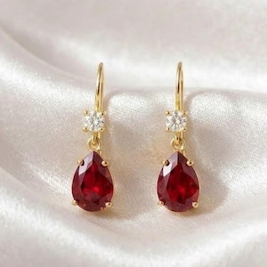 May include: A pair of gold-colored drop earrings. Each earring features a small, square, clear gemstone above a teardrop-shaped, deep red gemstone. The earrings hang from a gold-colored hook. The background is a white, silky fabric.