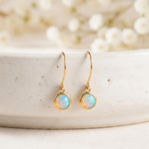 Pode incluir: Um par de brincos pendurados dourados com pedras de opala redondas e iridescentes. Os brincos têm um design simples e elegante, com fecho de gancho. Os opalas exibem uma mistura de tons azuis, brancos e amarelos. Os brincos são exibidos em uma superfície branca.