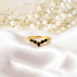 May include: A gold ring with a V-shaped design, set with several black, oval gemstones. The ring is displayed on white silk fabric, with a blurred background of white flowers. The ring's design is elegant and eye-catching.