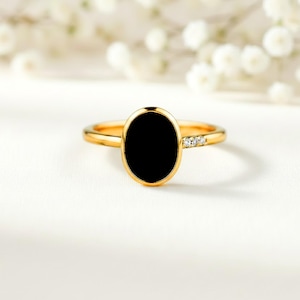Op de afbeelding: Een gouden ring met een ovale zwarte steen als middelpunt. De gouden band heeft kleine, heldere stenen. De ring staat tegen een witte achtergrond met zachte, vage witte bloemen. Een sieraad.