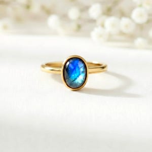 Puede incluir: Un anillo de oro con una piedra preciosa ovalada azul iridiscente. El anillo tiene una banda delgada y pulida, y la piedra preciosa está engastada en un bisel de oro. El anillo se muestra sobre una superficie blanca con iluminación suave y un fondo floral borroso.