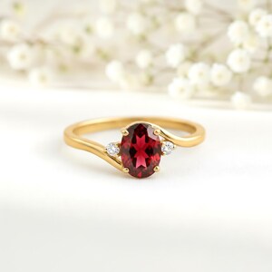 May include: A gold ring featuring an oval-cut, deep red gemstone, flanked by two smaller, round, clear gemstones. The ring's band has a delicate, flowing design. The background is a soft white with blurred white flowers.