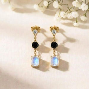 May include: A pair of gold earrings with a three-stone design. Each earring has a small, clear stone at the top, a black stone in the middle, and a rectangular, iridescent stone at the bottom. The earrings are set against a soft, neutral background.