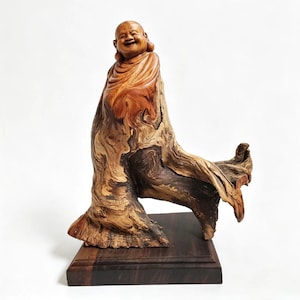 Puede incluir: Una escultura de madera de un Buda sonriente. La figura está tallada en un trozo de madera con una veta natural y fluida. La escultura está montada sobre una base de madera oscura.