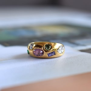 Könnte beinhalten: Ein goldener Bandring mit einer Vielzahl von bunten Edelsteinen, die in die Oberfläche eingelassen sind. Die Steine umfassen einen großen ovalen rosa Edelstein, einen runden olivgrünen Edelstein, einen rechteckigen violetten Stein und klare weiße Steine.