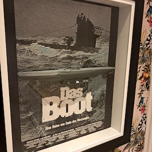 May include: Framed black and white movie poster for "Das Boot." The poster features a submarine and the film's title in large letters. The frame is black with a white mat, mounted on a floral wallpaper background.