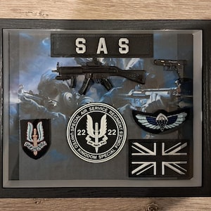 May include: Framed display with SAS patches, a submachine gun, and a handgun. The display includes a Union Jack patch, a winged emblem, and a patch with the text "SAS". The background shows soldiers in combat.