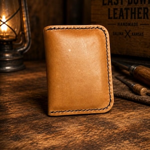 May include: A tan leather card holder featuring rounded corners and thick black saddle stitching along the edges. The surface of the leather has a smooth, matte finish with natural variations in color and texture throughout the material.