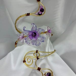 May include: Gold-toned wire jewelry with floral and spiral designs. The jewelry features purple flower accents and small purple beads. The wire is wrapped with brown wire in certain areas. The jewelry is displayed on a white satin background.