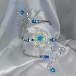 Peut inclure: Un bracelet en fil d'argent avec un motif floral. Le bracelet présente une grande fleur translucide avec des accents bleus au centre, entourée de petites fleurs bleues et de volutes décoratives. La pièce est posée sur un fond blanc satiné.
