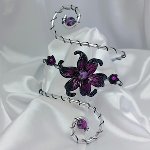 May include: An ornate arm cuff in silver metal, featuring a central dark purple flower and smaller buds. The design incorporates spiral elements and spherical purple beads. The cuff is designed to be worn on the upper arm.
