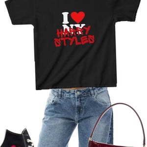 May include: A black t-shirt with white and red text reading I heart NY HARRY STYLES, blue denim jeans, black high-top sneakers with a red heart logo, a dark red shoulder bag, and black rectangular sunglasses arranged in a flat lay style.