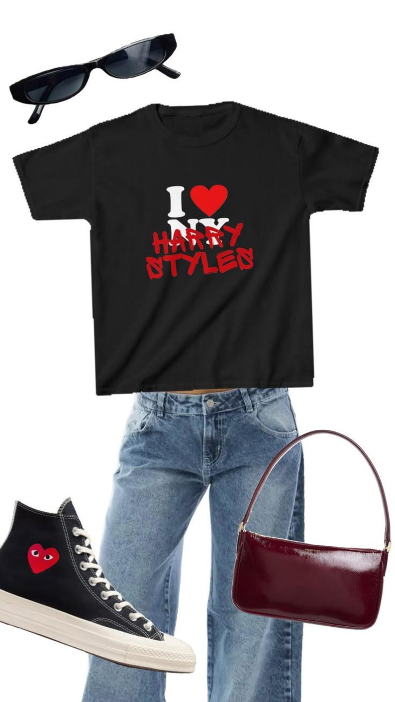 May include: A black t-shirt with white and red text reading I heart NY HARRY STYLES, blue denim jeans, black high-top sneakers with a red heart logo, a dark red shoulder bag, and black rectangular sunglasses arranged in a flat lay style.
