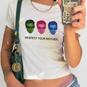 Puede incluir: Una camiseta blanca de manga corta con un gráfico de tres rostros idénticos con gafas de sol en verde, rosa y azul. Debajo de los rostros, el texto dice RESPECT YOUR MOTHER. La camiseta se combina con un bolso de hombro verde oscuro con un dije dorado de sol y luna.