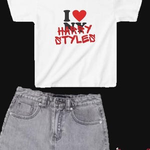 May include: A white short-sleeve t-shirt with the text I heart NY HARRY STYLES in black and red, a light wash denim mini skirt, red oval-frame sunglasses, and low-top sneakers in a two-tone white and dark red colorway with a side swoosh.