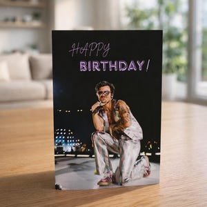 May include: A greeting card with a black background and the words "HAPPY BIRTHDAY!" in a neon-style font. The card features a photo of a person kneeling, wearing a striped outfit and sunglasses.