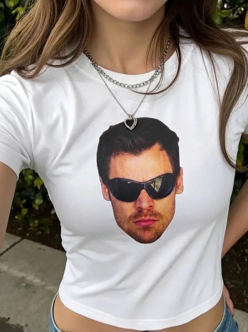 May include: A white short-sleeved t-shirt featuring a large, centered graphic print of a man's face wearing dark, wraparound sunglasses. The shirt is paired with a layered silver-toned chain necklace that includes a small heart-shaped pendant.